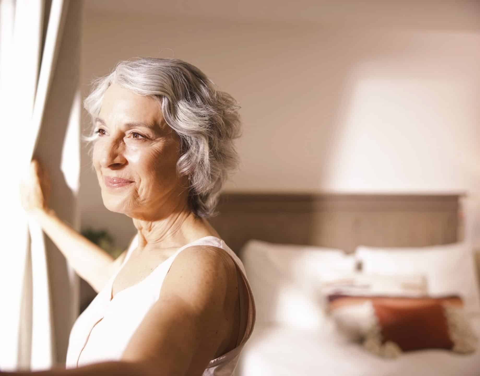 An older woman with short gray hair stands by a window, gently holding open white curtains. Sunlight shines on her face, and there is a neatly made bed in the softly lit background.