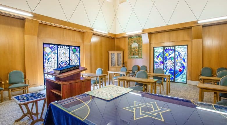A bright synagogue room radiating spirituality, with wooden walls, green chairs, tables, stained glass windows, and a bimah draped with a cloth featuring a Star of David. Natural light streams in from above.