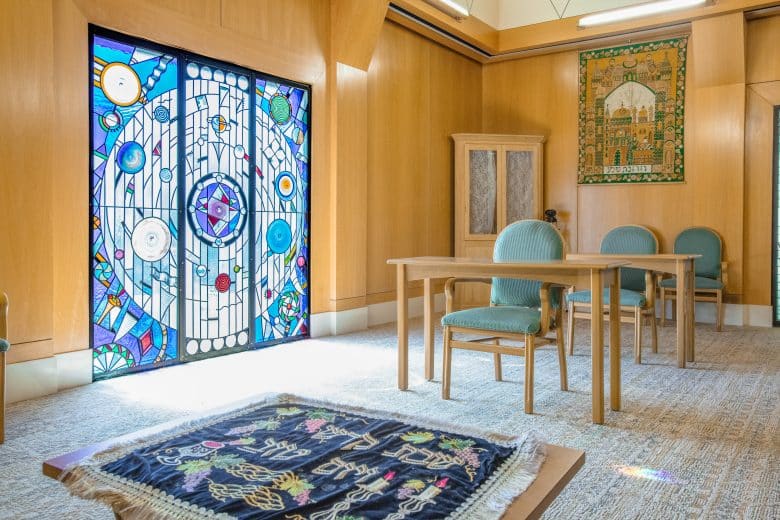 A bright room with light wood walls, green upholstered chairs, a wooden table, a cabinet, and a colorful stained glass window with Jewish symbols creates an atmosphere of spirituality. An embroidered cloth with Hebrew designs rests on the floor.