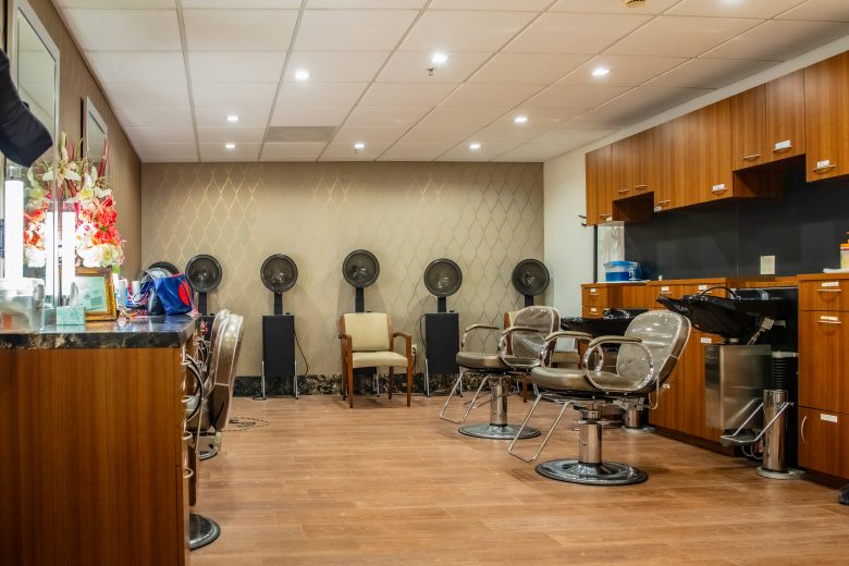 A modern hair salon interior with brown wooden floors, salon chairs, wash sinks, and hairdryers in the back row creates a chic lifestyle vibe. Cabinets line the right wall, while decorative flowers brighten the counter to the left. The Reutlinger Amenities and Services