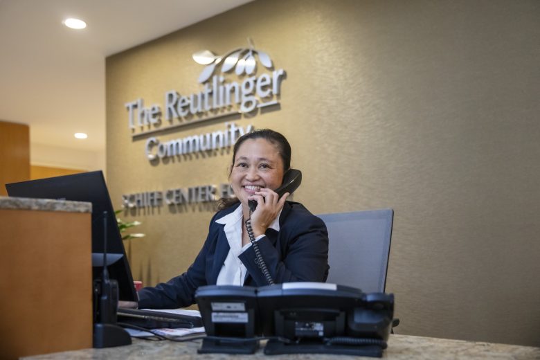 A woman in a suit sits at a reception desk, smiling and talking on the phone in front of a sign that reads The Reutlinger Community. A computer monitor and office phone are on the desk.