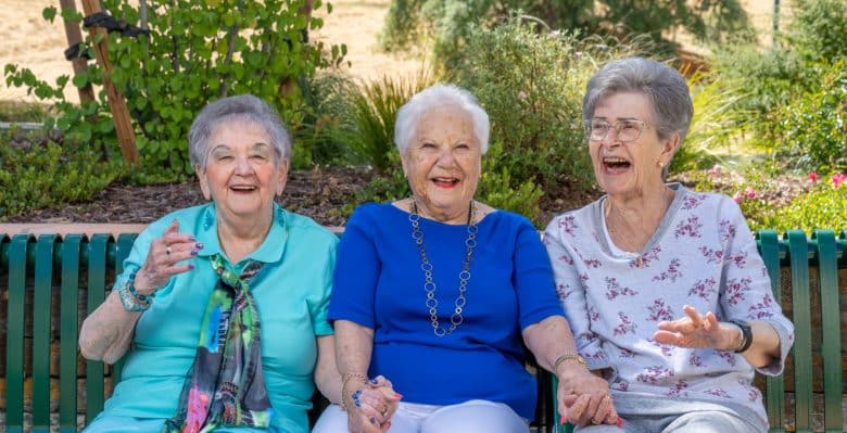 Three elderly women sit on a green bench outdoors, smiling and laughing together as they discuss living options. The background features greenery and trees, suggesting a peaceful park or garden setting.
