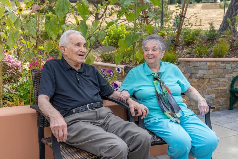 Two elderly people sit holding hands and smiling on patio chairs in a garden, surrounded by greenery and flowers, enjoying a sunny day outdoors while exploring different living options.
