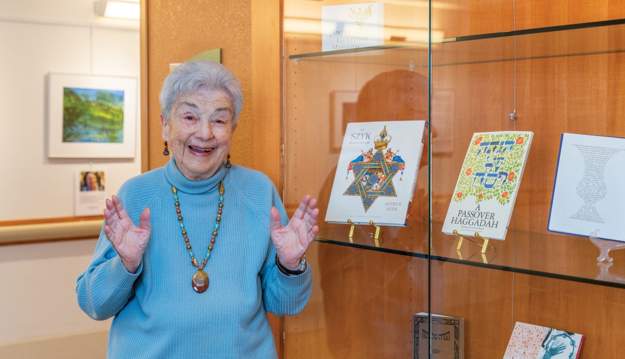 An elderly woman in a blue sweater and beaded necklace smiles and gestures with raised hands, standing beside a glass display case containing illustrated books and documents that reflect her deep sense of spirituality.