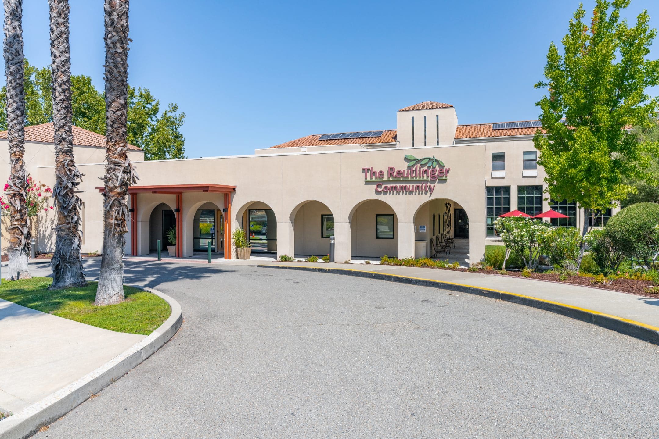 A beige stucco building with arched entryways, “The Reutlinger Community” sign, palm trees, and solar panels on the roof sits on a sunny day with a curved driveway in front—perfect for your next brochure or to download for more information.