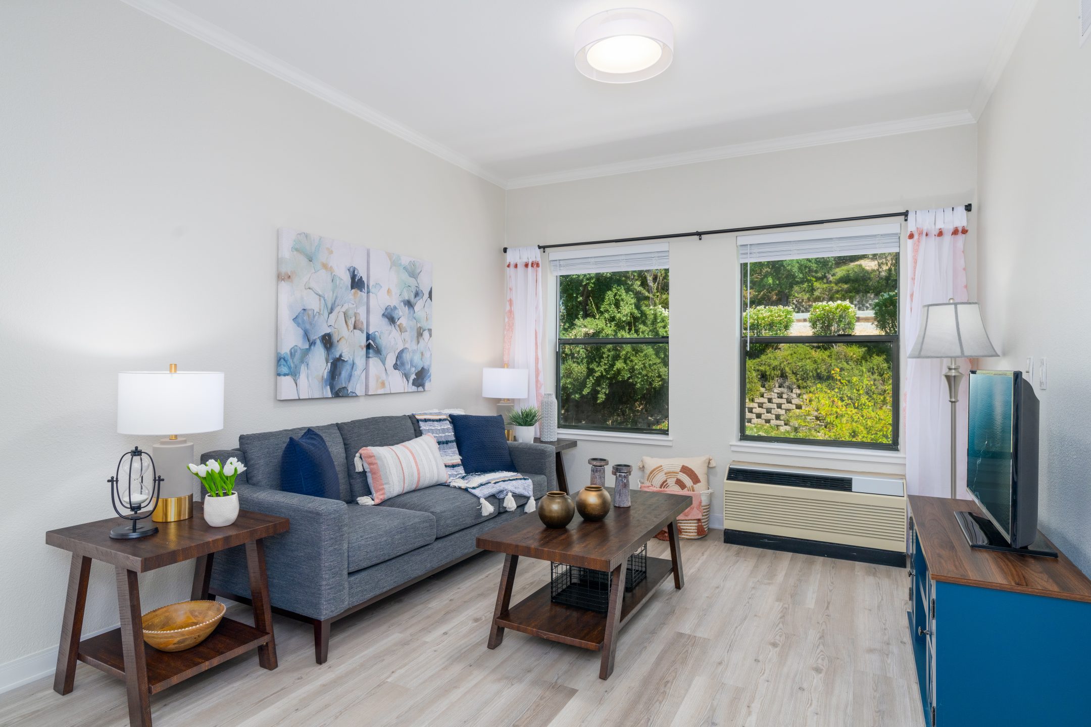 Modern living room with a gray sofa, two wooden side tables with lamps, a coffee table, TV stand, and wall art. Large windows reveal greenery outside. Ideal for short-term stays, light wood floors and white walls brighten the inviting space.