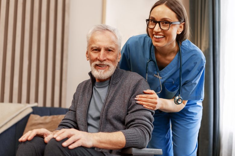 An older man with gray hair and a beard sits in a chair, smiling, while a young nurse in blue scrubs and glasses stands beside him, also smiling and supportively holding his arm at The Reutlinger living options.