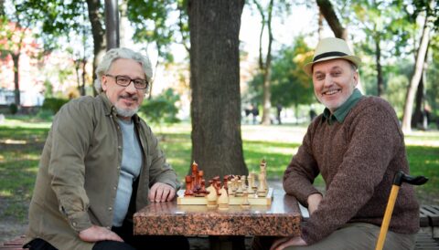 Two older men sit at a park table playing chess, smiling at the camera. Surrounded by trees and greenery, one man—enjoying The Reutlinger living options—wears a hat and holds a cane on this bright, sunny day.