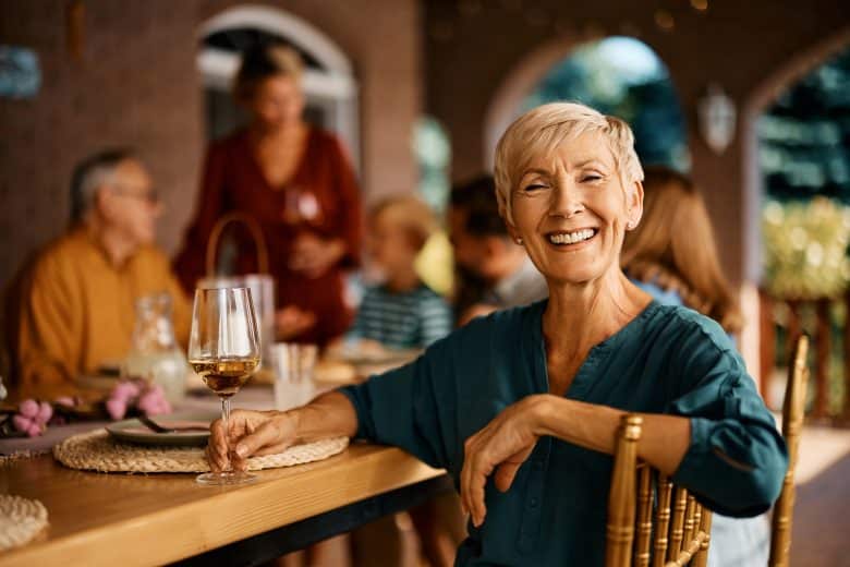 A smiling older woman with short blond hair sits at an outdoor table holding a glass of white wine, with a group of people gathered and talking in the background.