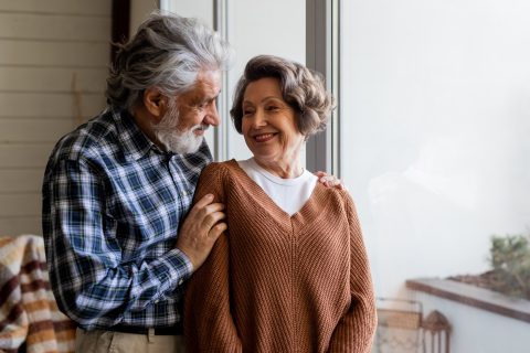 An elderly couple stands by a large window, smiling warmly at each other in their cozy, sunlit room—reflecting the happiness and contentment offered by The Reutlinger living options.