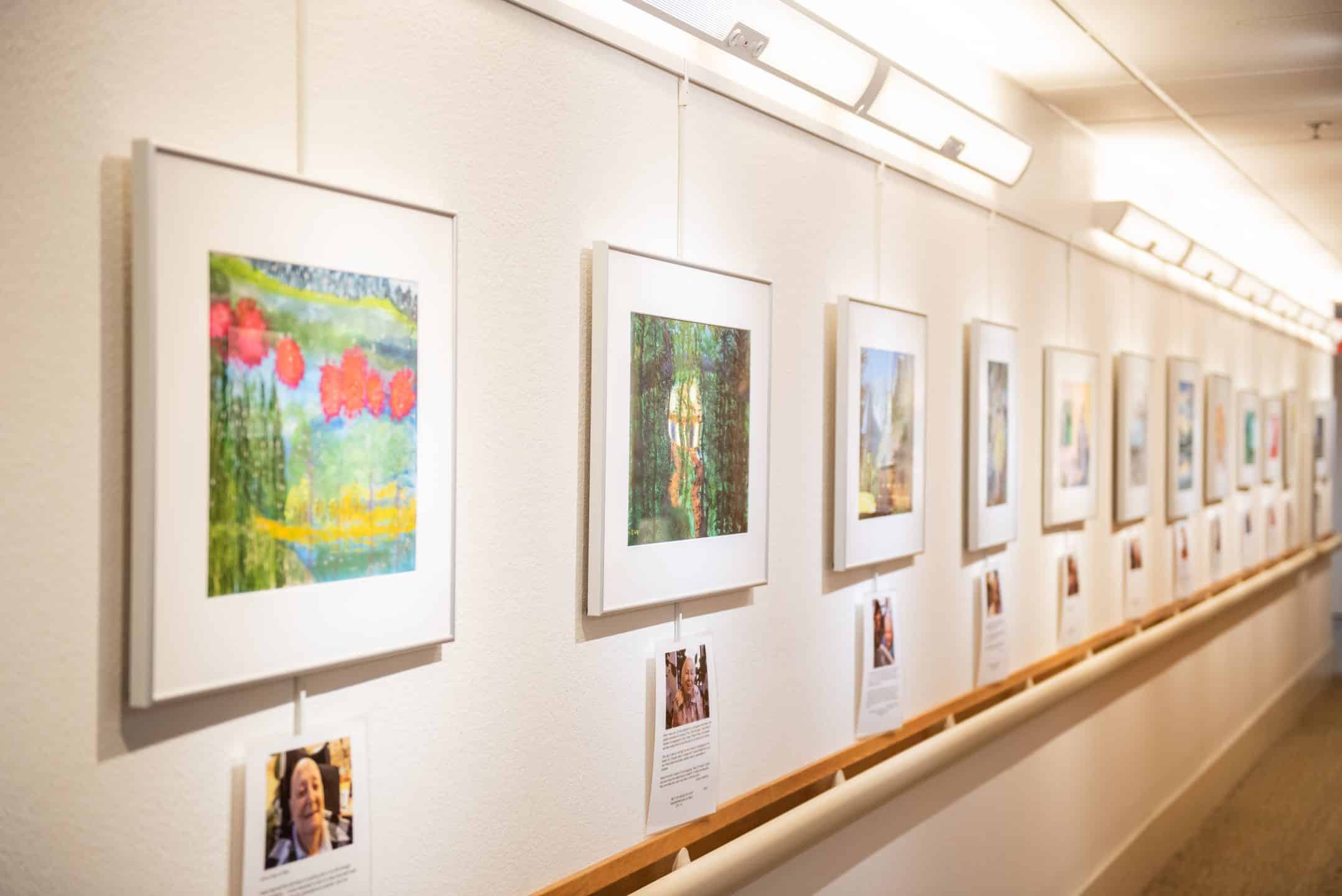 Framed artwork hangs in a well-lit hallway gallery. Each piece is accompanied by a printed description or biography below, displayed in a single row along the white wall.