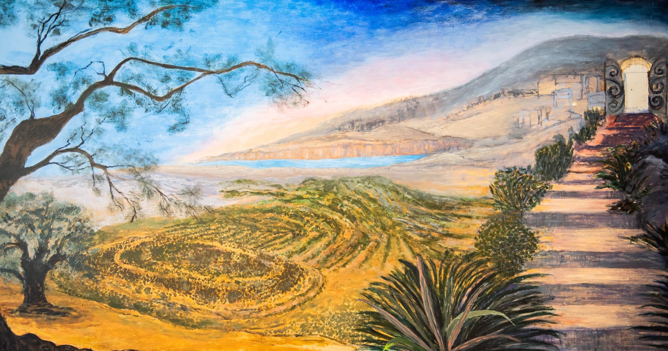 A painted landscape featuring rolling fields, a winding path lined with plants, a tree on the left, distant mountains, a body of water, and a gate leading to a building on a sunlit hillside under a blue sky.