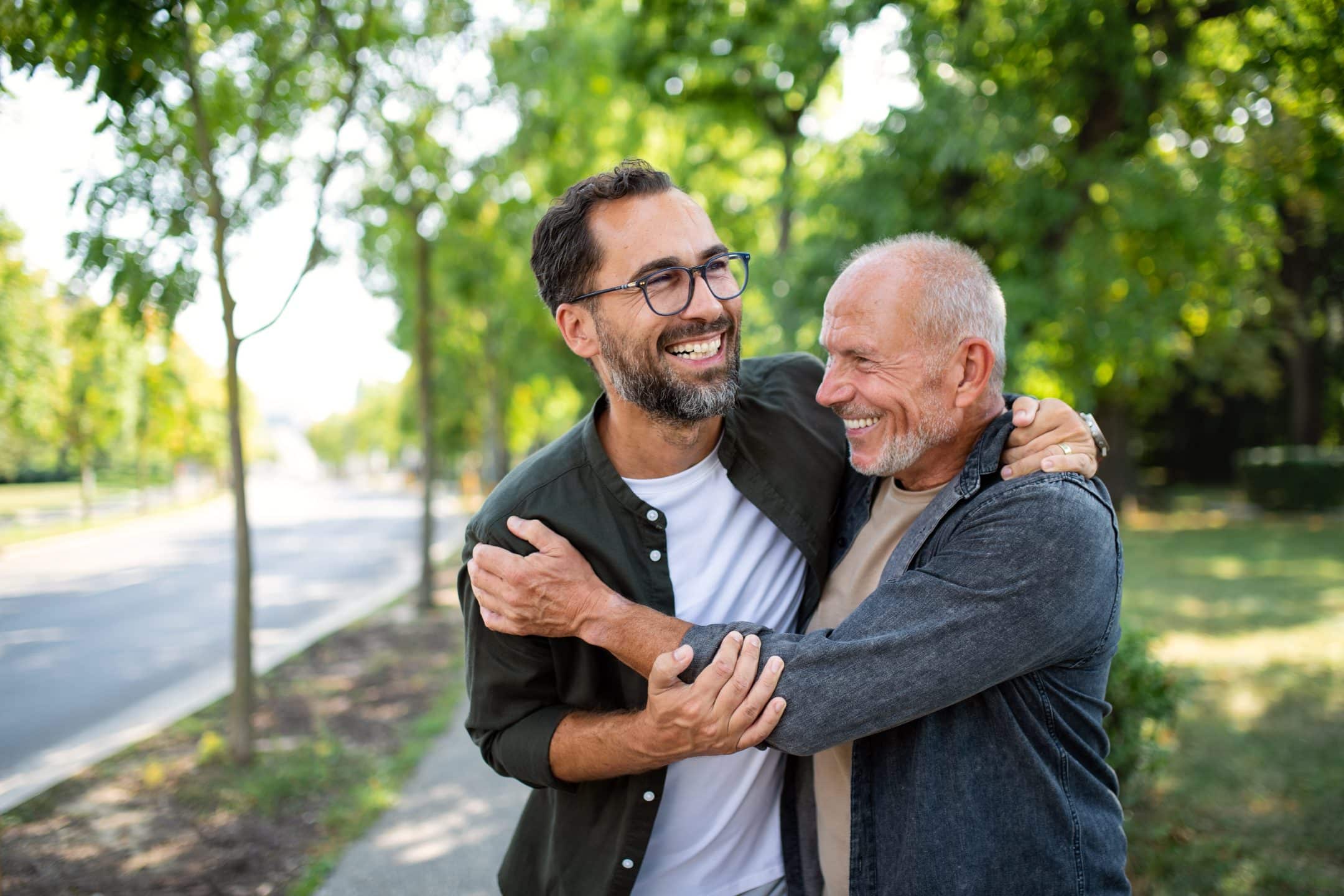 Two smiling men, one older and one younger, stand outdoors on a sunny day, hugging and laughing together on a tree-lined sidewalk. For more moments like this, contact us today.