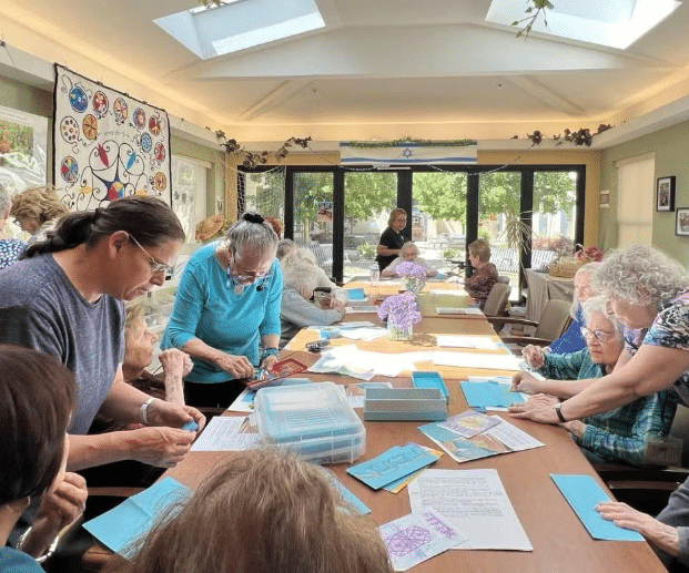 A group of elderly women are seated around a large table, enhancing their resident experience as they engage in a crafting activity with paper. The bright room, filled with natural light, is decorated with colorful art and flowers.