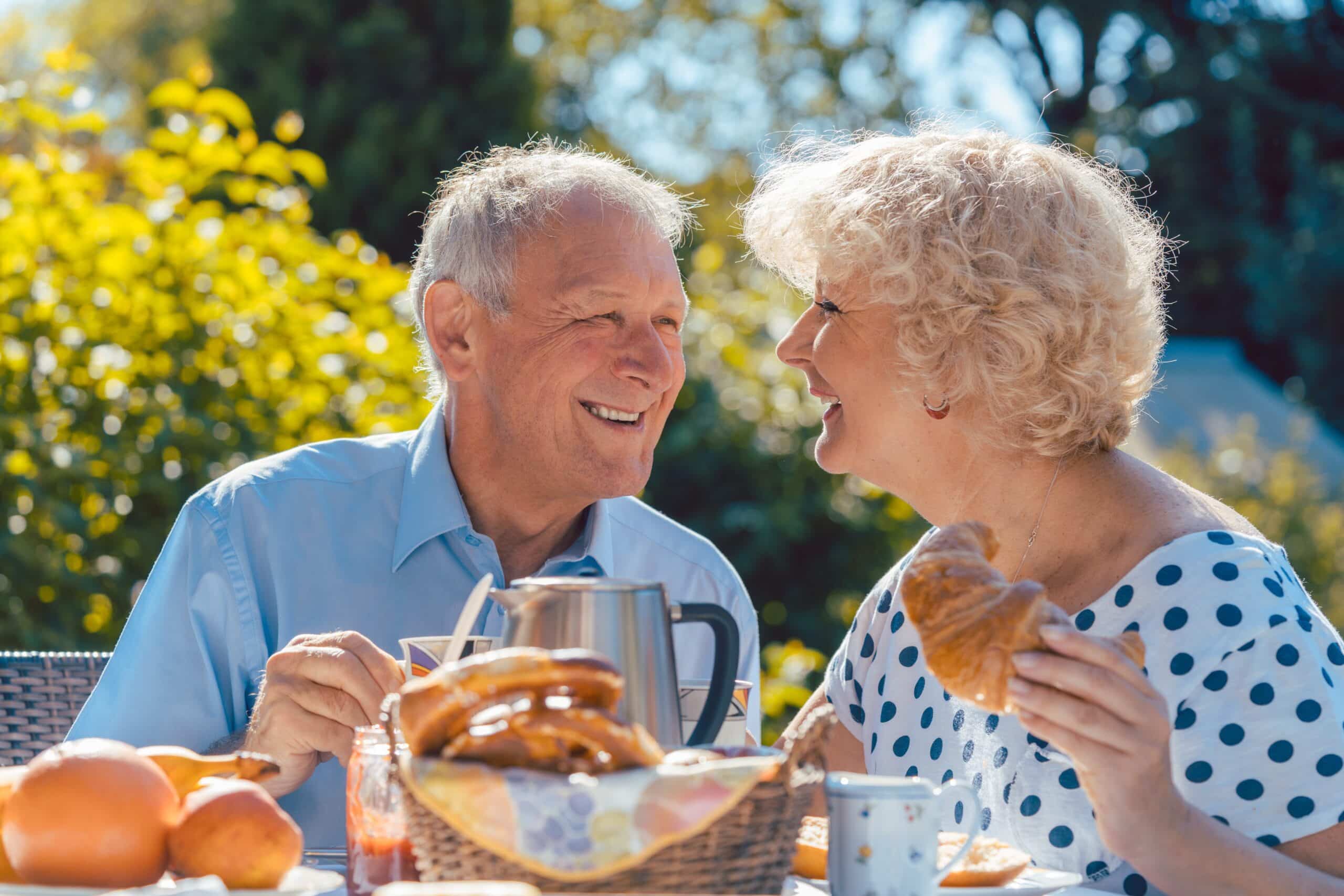 An older couple sits at an outdoor table, smiling at each other while enjoying a Brunch and Learn with croissants, pastries, fruit, and coffee on a sunny day.