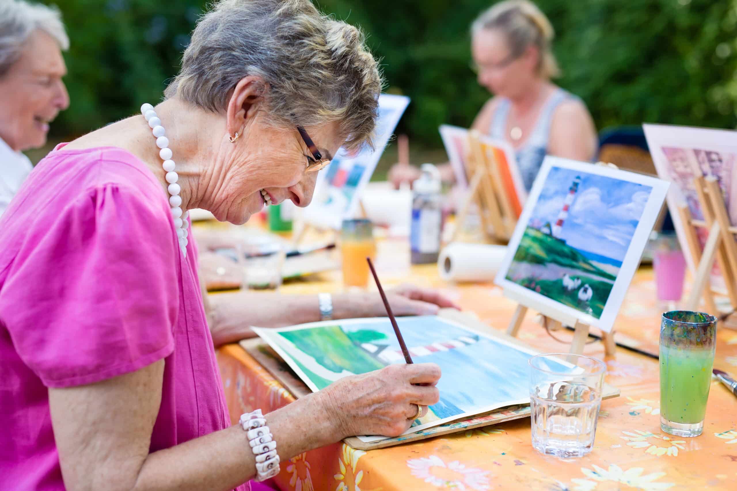 An elderly woman in a pink shirt smiles while painting a landscape at an outdoor Paint and Sip class with other seniors. She sits at a table with art supplies, a glass of water, and her finished painting of a lighthouse nearby.